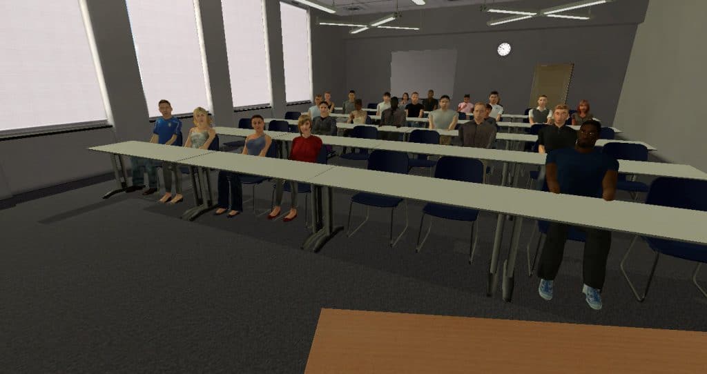 A classroom half full. Tables extend the width of the room. You are standing behind a desk presenting, giving a feeling of security. The audience is mostly attentive, though two have mobile phones out.